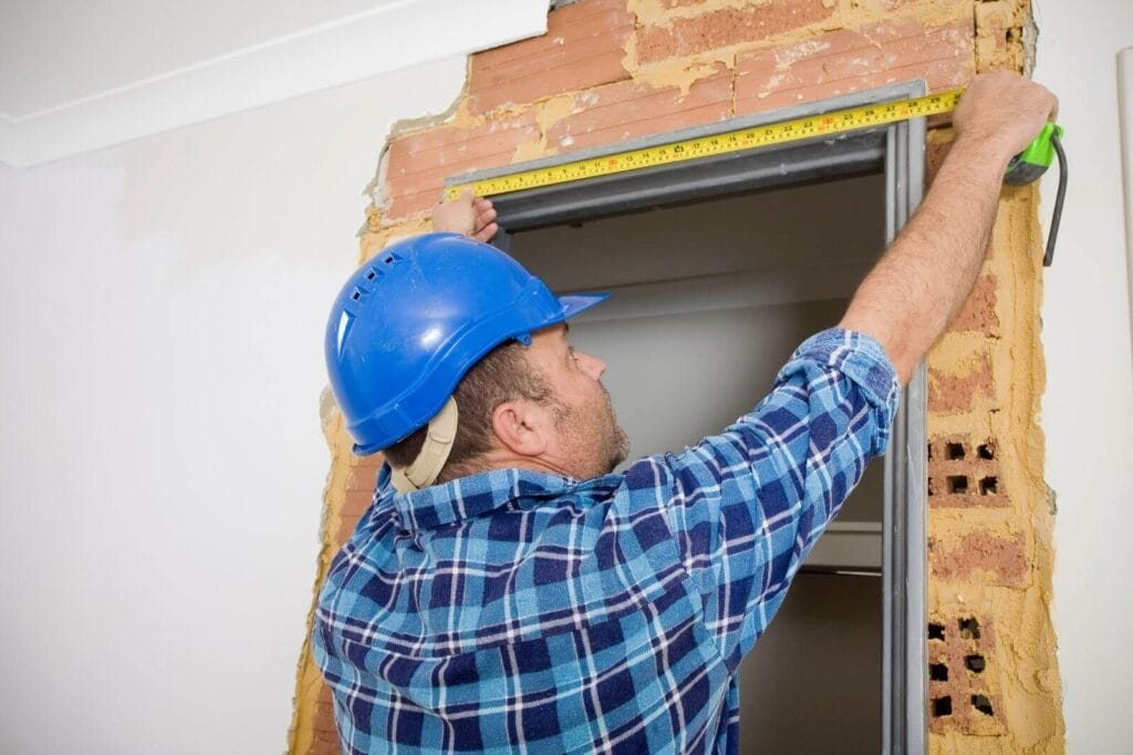 A man wearing a hard hat measures the width of a doorway.