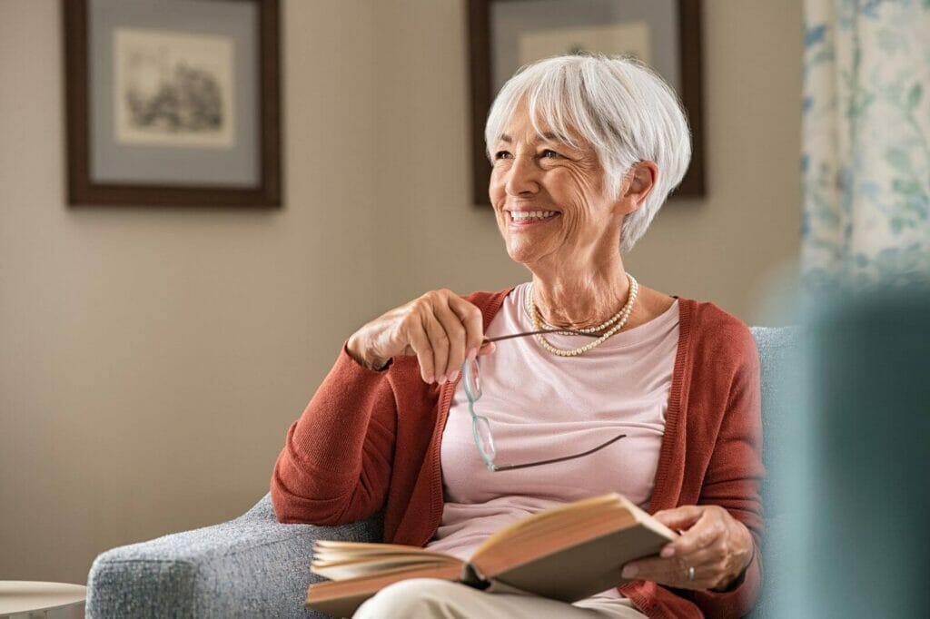 A smiling older adult woman sits in an armchair holding a book and a pair of glasses.