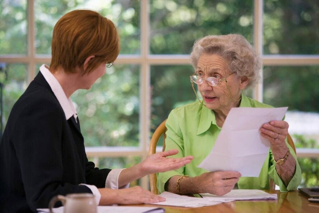 An older adult woman wearing reading glasses and holding a piece of paper sits at a table and talks with a younger woman.