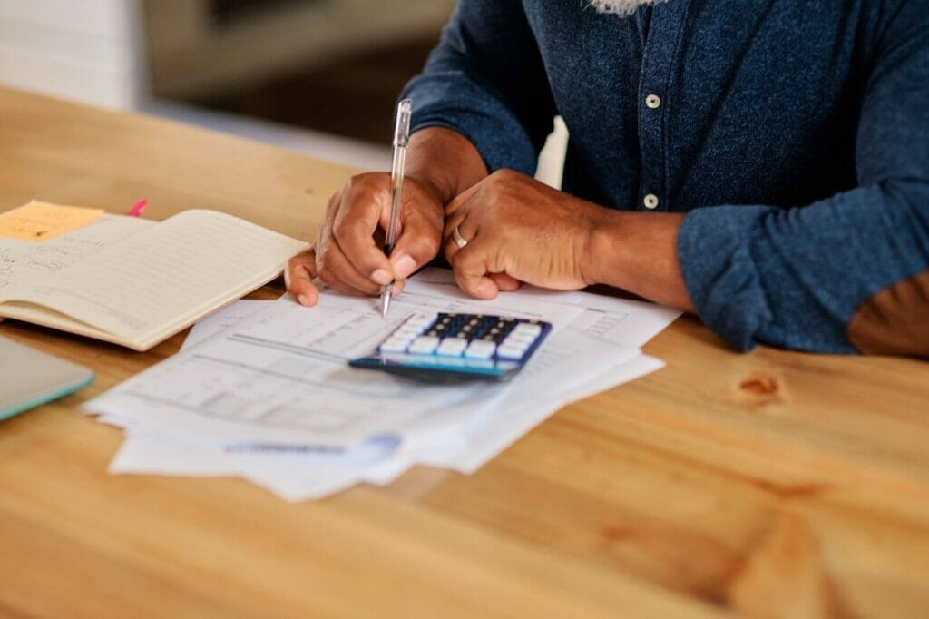 An adult man's hands are shown holding a pen and filling out paperwork. There is a calculator on the desk.