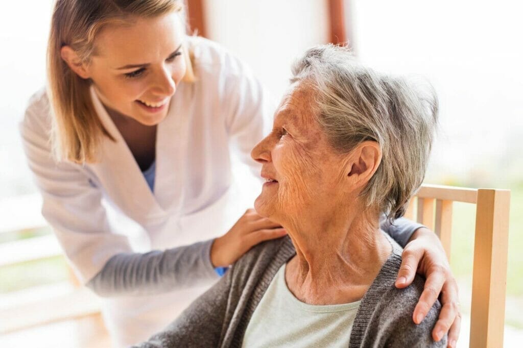 An adult woman caregiver has her arms around a seated older adult woman. They smile at each other.