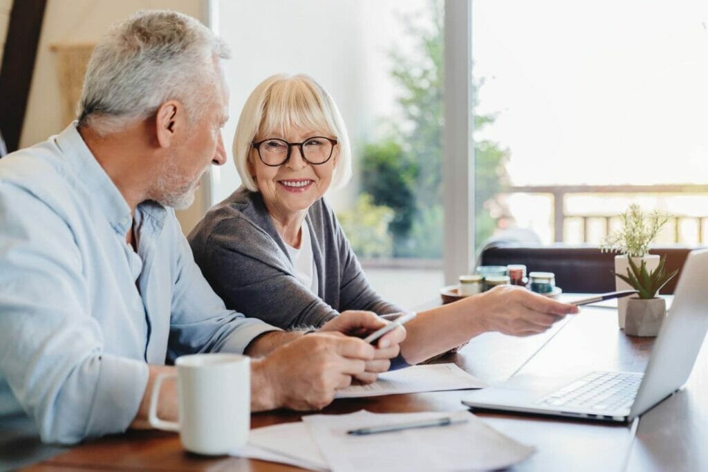 An older adult man and woman sit at a table with a laptop and paperwork. They smile at each other.