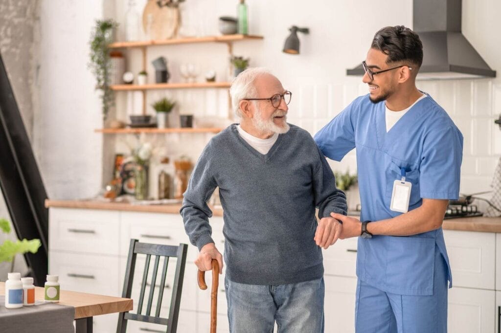 An adult man wearing scrubs assists an older adult man using a cane.