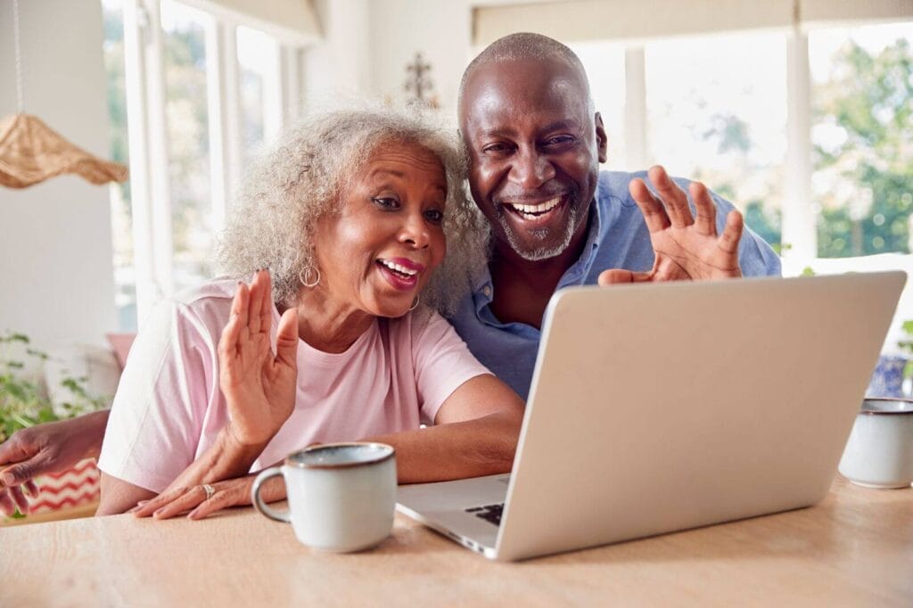 An older adult man and woman sit at a table, smiling and waving at their laptop.