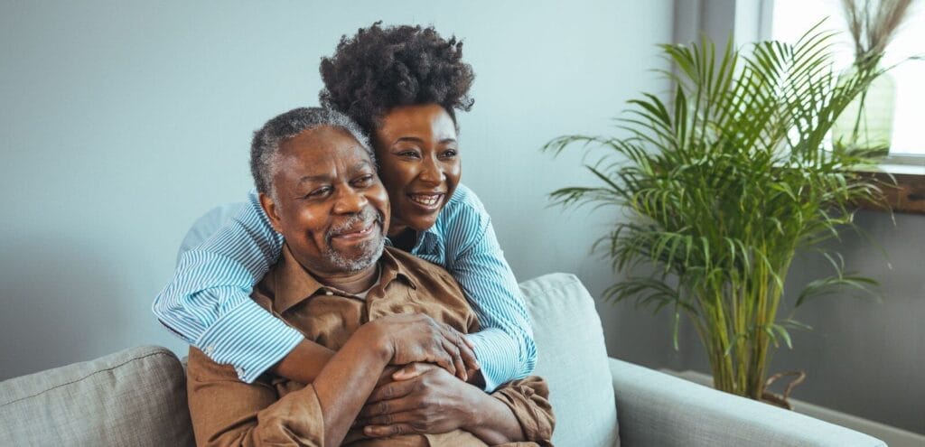 A woman stands behind an older adult man, hugging him. They both smile.