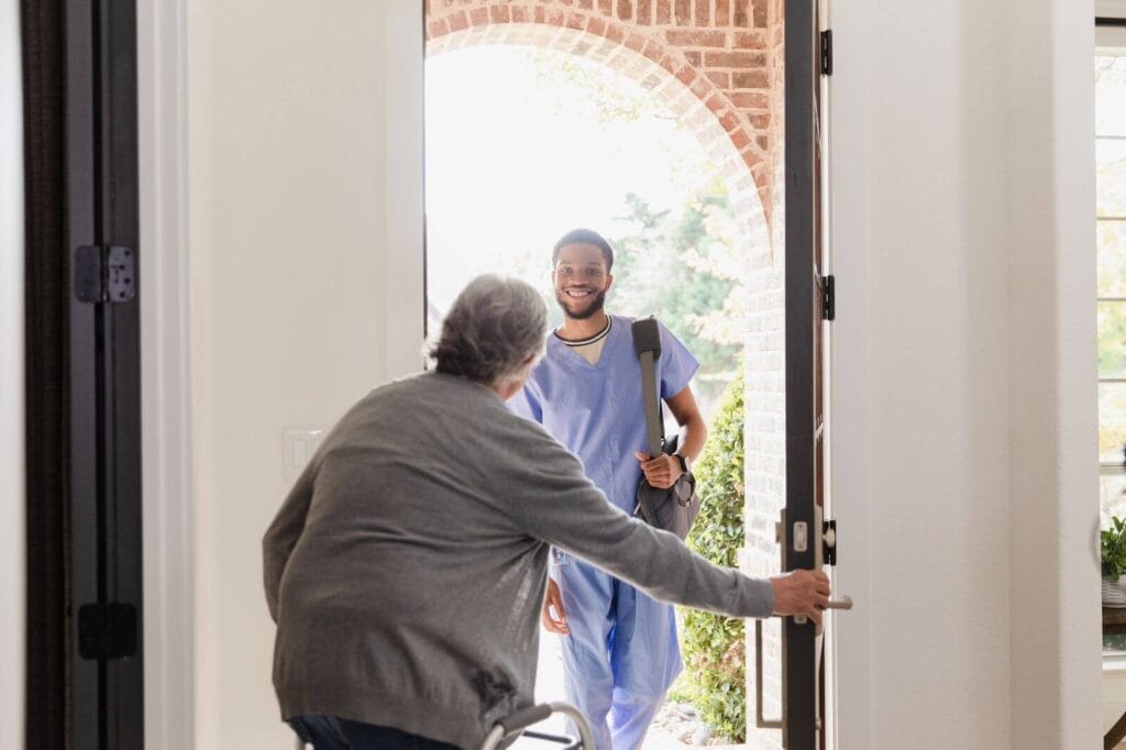 An older adult using a walker opens their front door to let in a smiling man wearing scrubs.