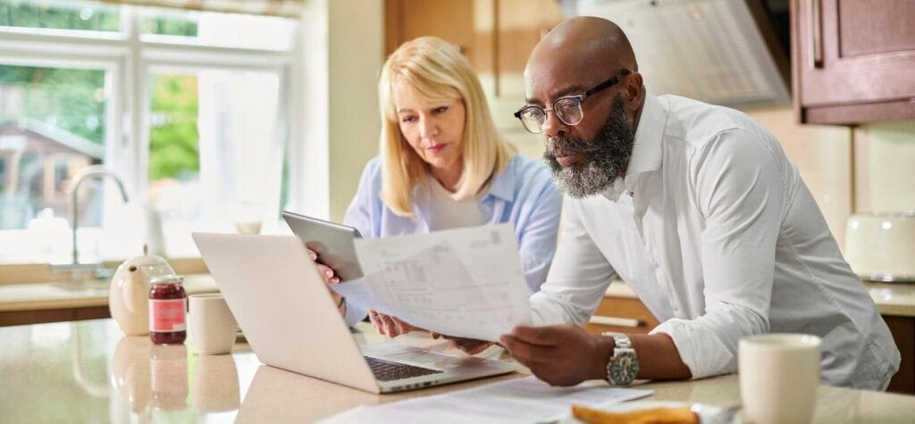 An older adult man and woman stand at a kitchen counter, looking over paperwork.