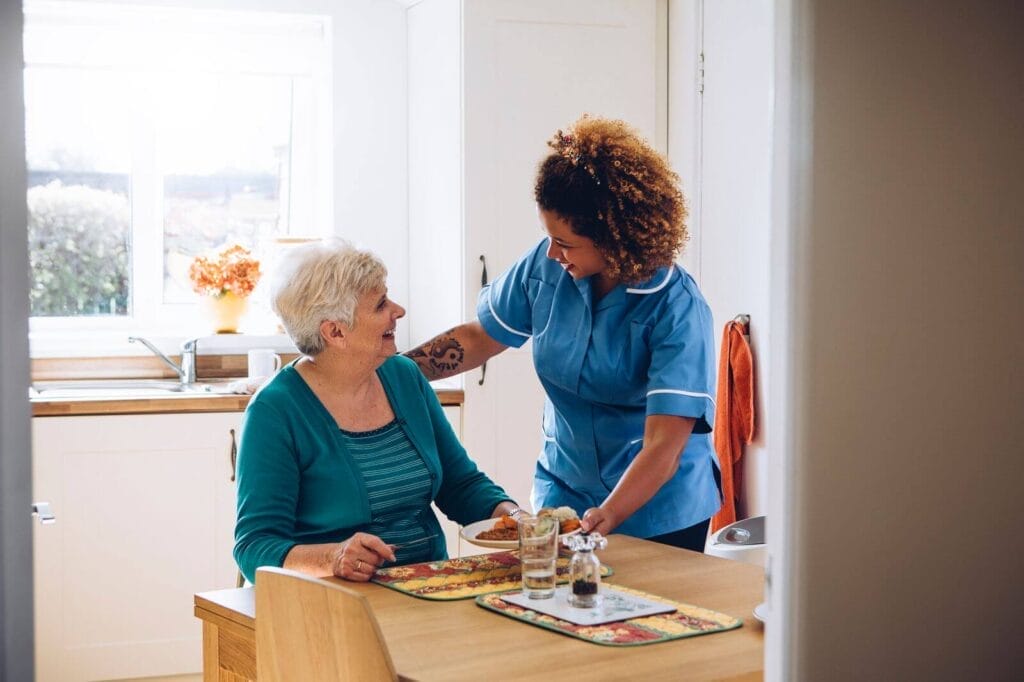 A female caregiver sets a plate of food in front of an older adult woman sitting at her kitchen table.