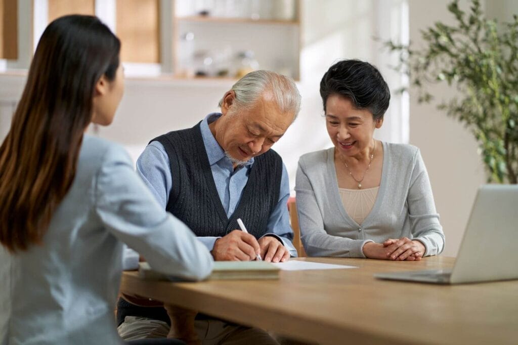An older adult man and woman sit at a table with a younger woman. They are signing paperwork.