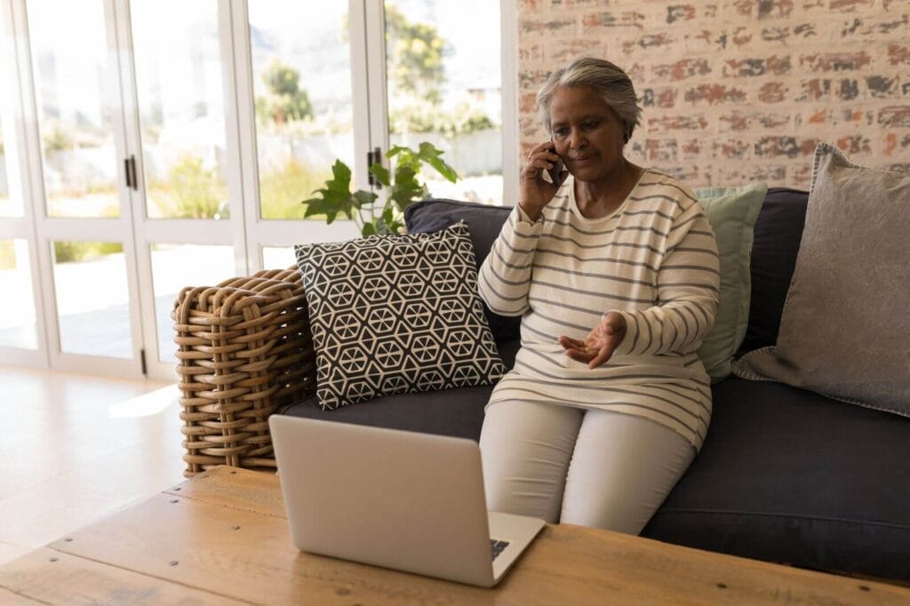 An older adult woman sits on a couch talking into her cell phone. A laptop sits on a coffee table in front of her.