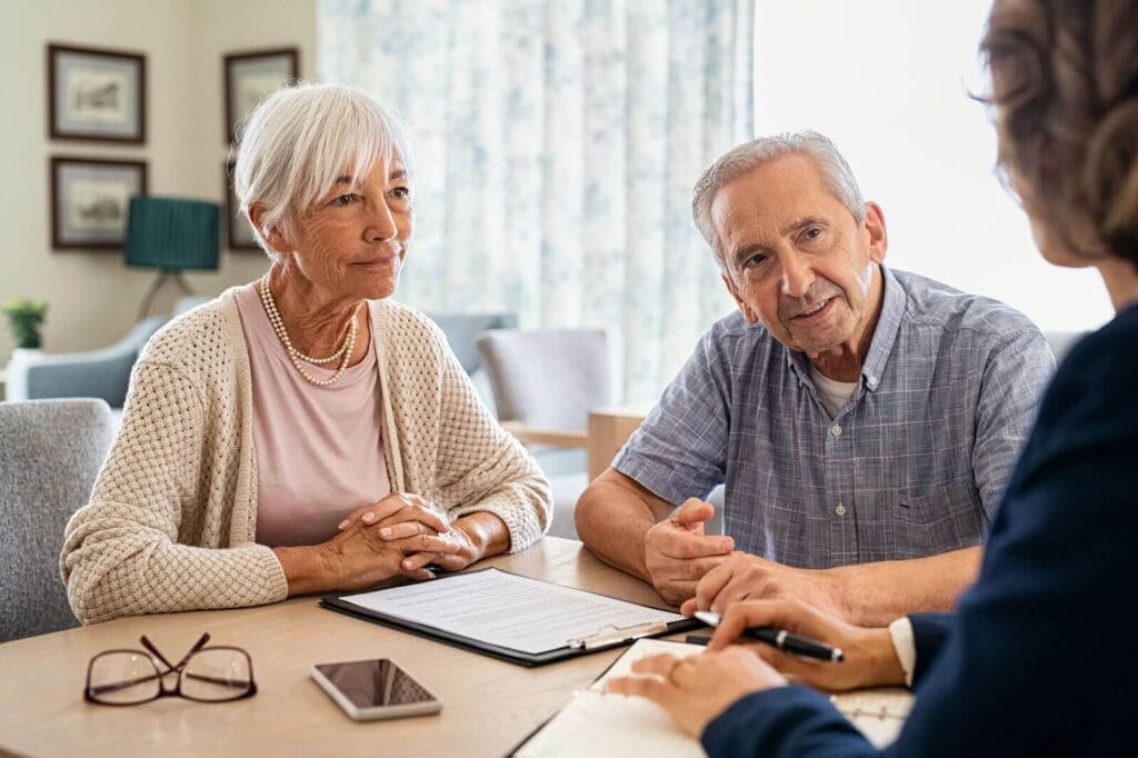 An older adult man and woman sit at a desk with a woman. They are going over paperwork.