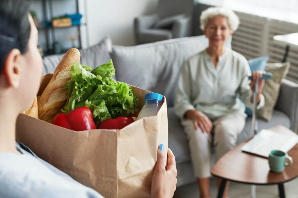 A woman carries a bag of groceries toward an older adult woman sitting on a couch.
