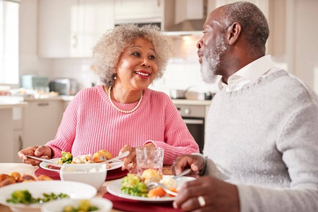 An older adult man and woman sit at their kitchen table eating a meal.