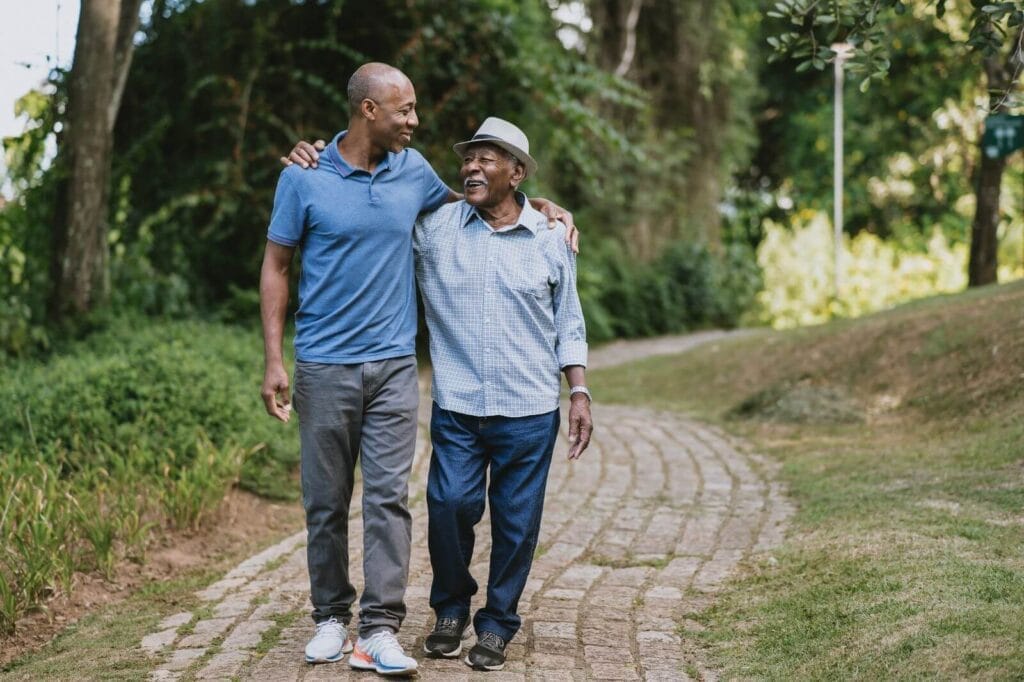 A man and an older adult man walk along a path with their arms around each other.