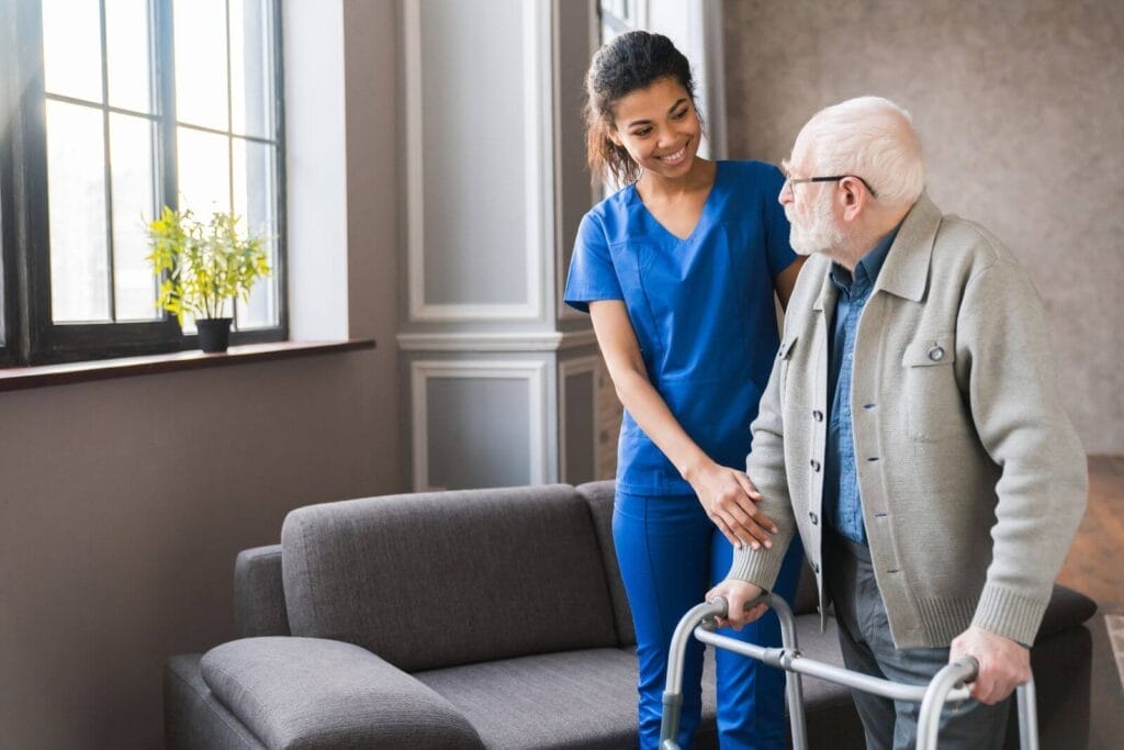 A woman wearing scrubs assists an older adult man using a walker.