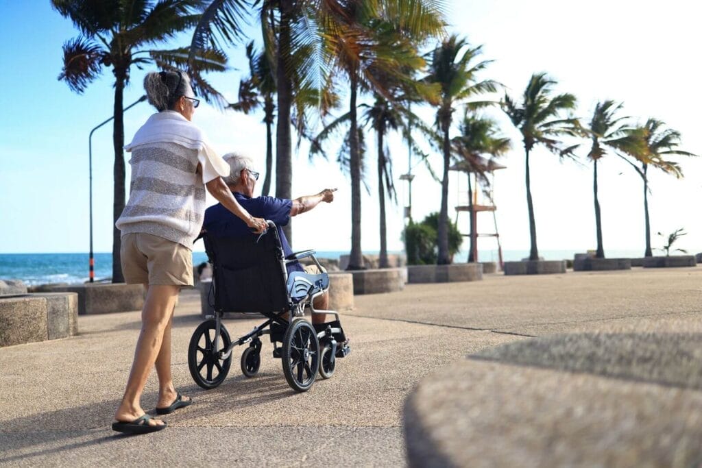 An older adult woman pushes an older adult man in a wheelchair down a walkway next to a beach with palm trees.