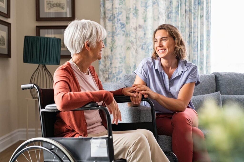 A woman sitting on a couch smiles and holds the hand of an older adult woman in a wheelchair.