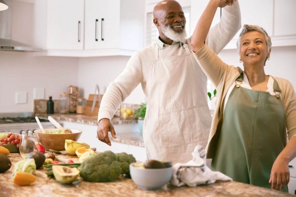 An older adult man and woman dance together in the kitchen as they prepare a meal.