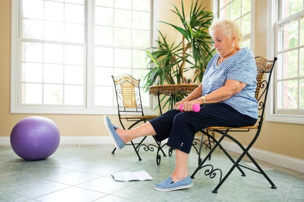 An older adult woman sits in a chair holding dumbbells with her right leg extended before her.