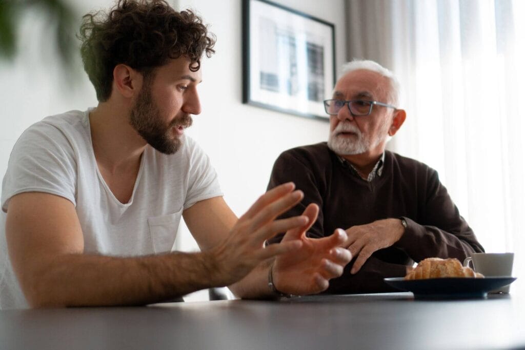 A man and an older adult man sit at a table talking.