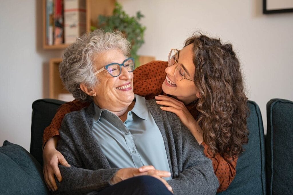 An older adult woman sits on a couch while a younger woman hugs her from behind.