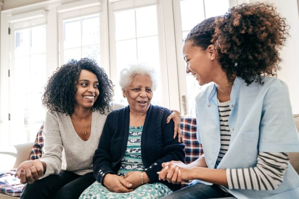 Two women sit on a couch on either side of an older adult woman. They are all smiling.