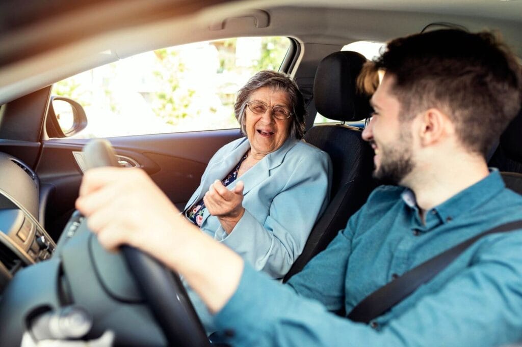 A man is driving a car, smiling at his older adult woman passenger.