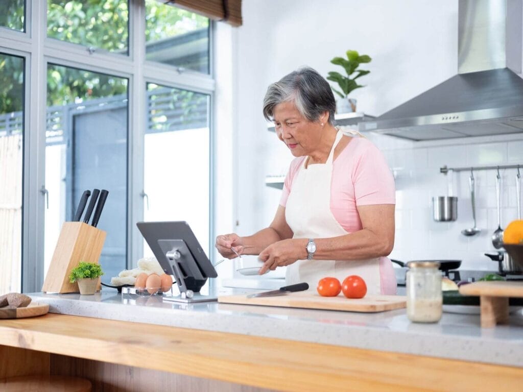 An older adult woman stands at her kitchen counter preparing a meal as she looks at her tablet.
