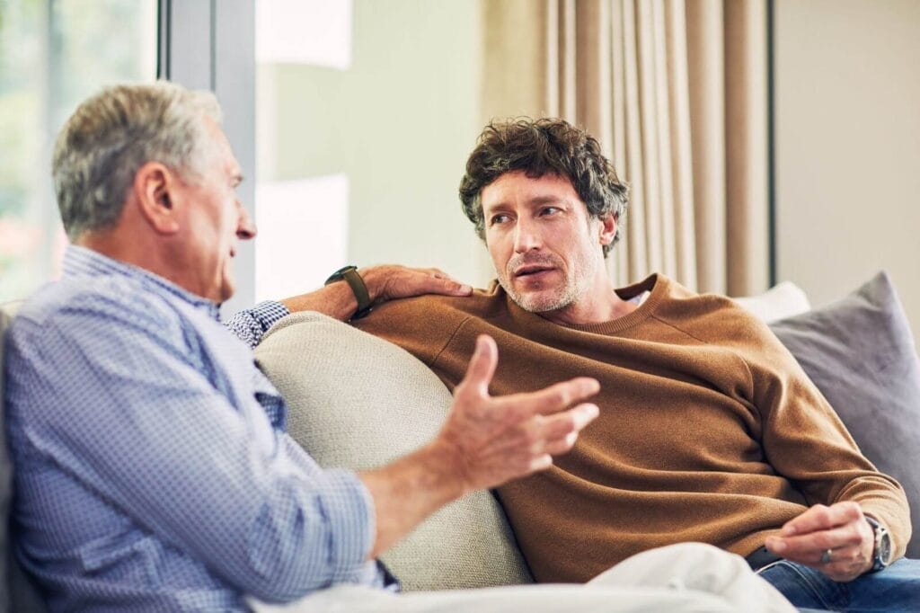 A man sits on the couch talking with an older adult man.