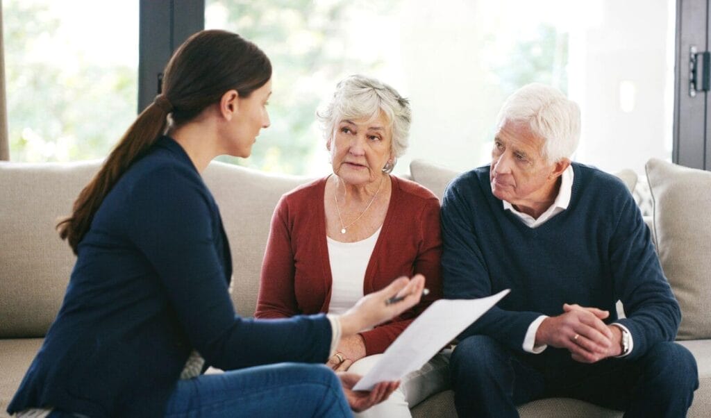 An older adult man and woman sit on a couch talking to a woman who is showing them some paperwork.