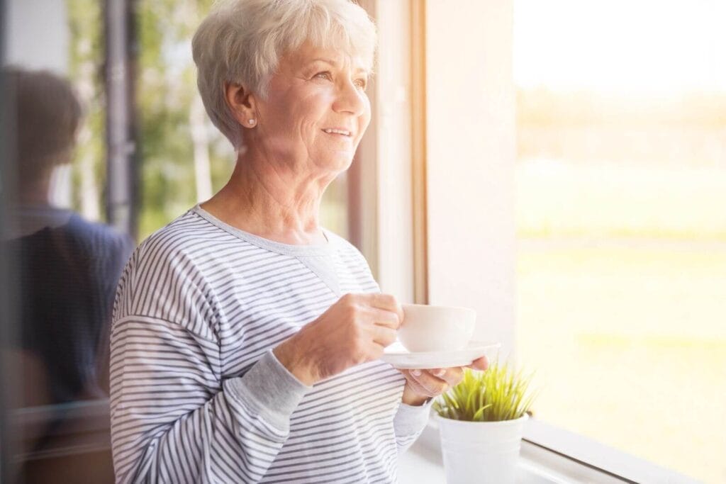 An older adult woman drinking coffee or tea looks out a window in the morning.