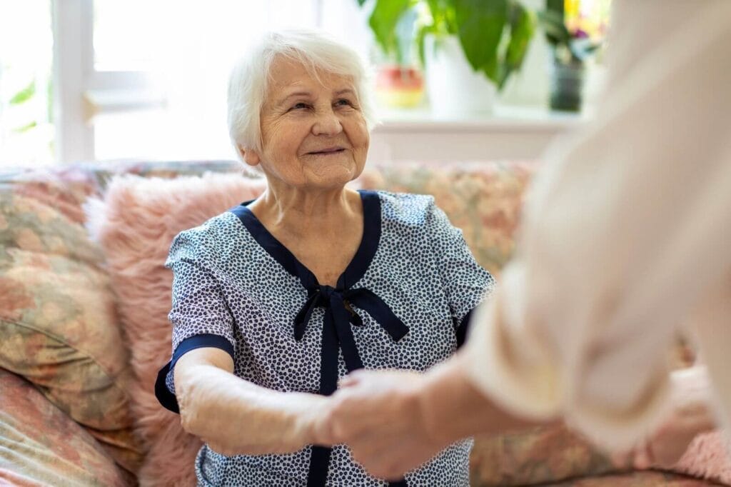 An older adult woman is sitting on a couch holding the hands of someone who is out of the frame.