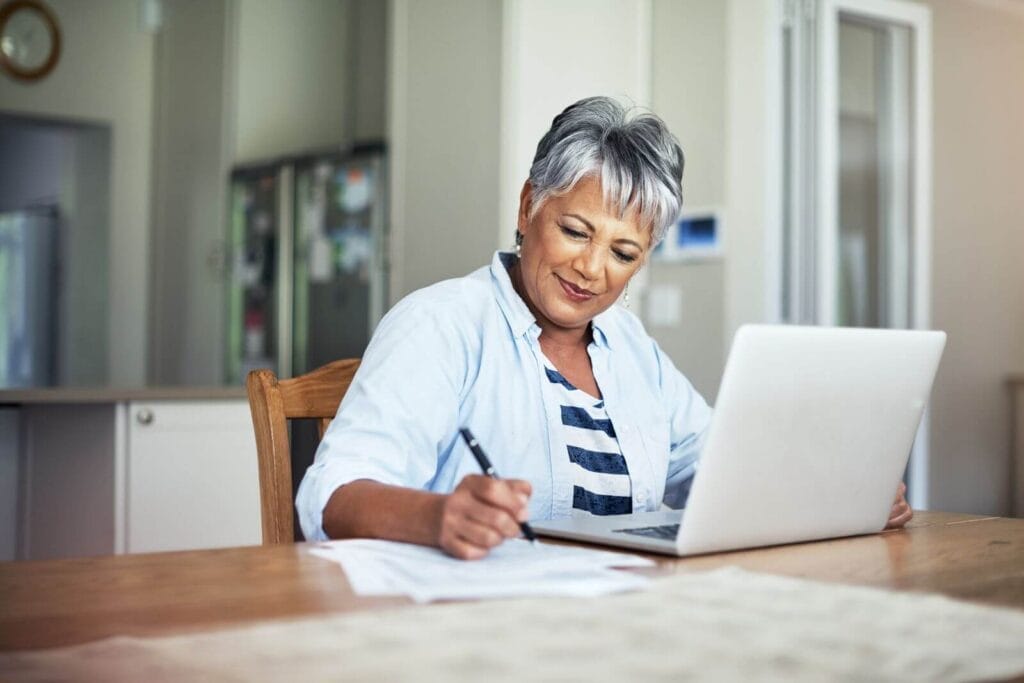 An older adult woman sits at a table in front of a laptop, writing on paper.