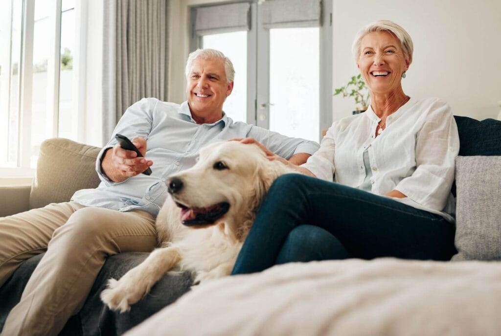 An older adult man and woman sit on a couch with their dog. The man is holding a remote control for a TV.