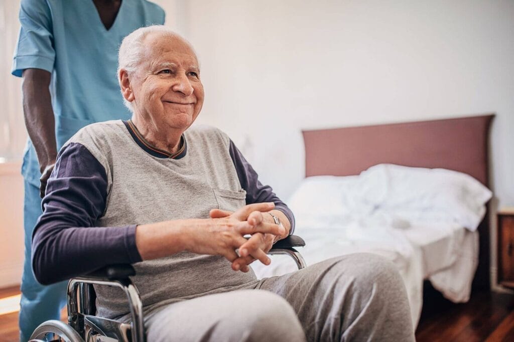 A male health care professional pushes an older adult man in a wheelchair out of a bedroom.