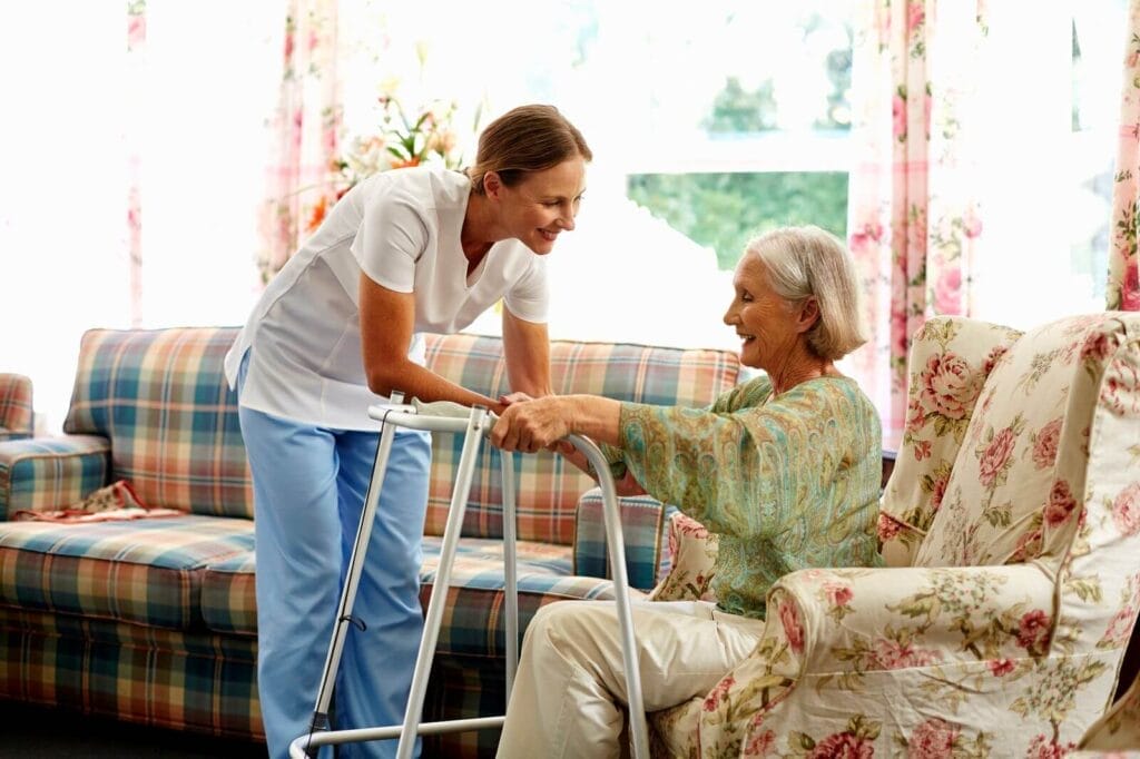 A female health care professional helps an older adult woman stand up using a walker.