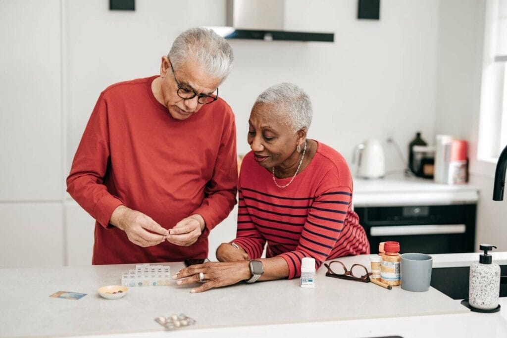 An older adult man and woman organize medication in a pillbox at their kitchen counter.