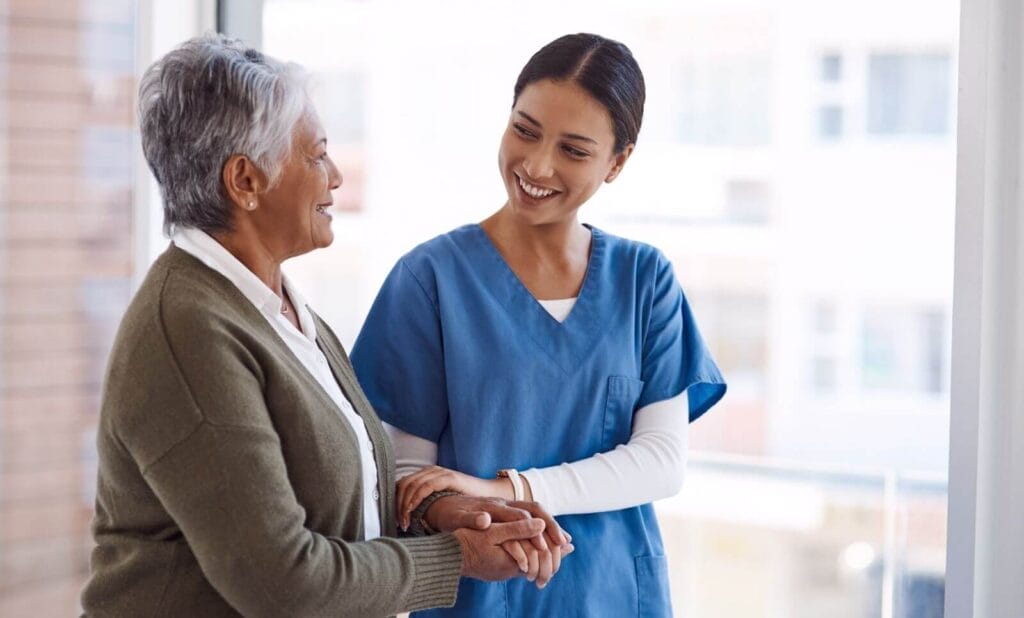A woman wearing scrubs holds the hands of an older adult woman. They are standing.