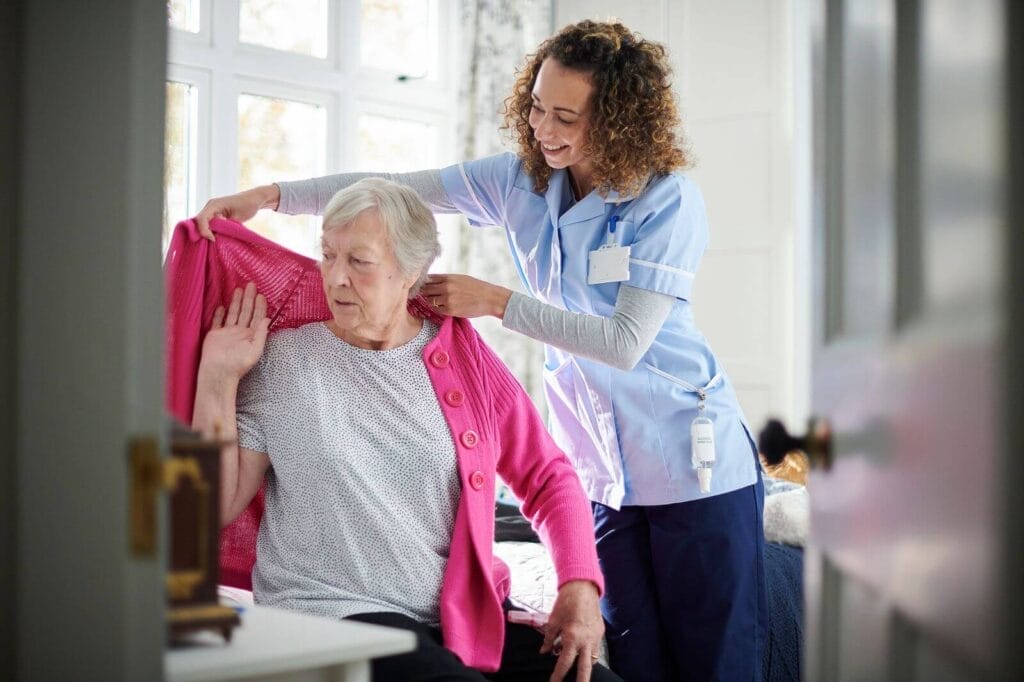 A female caregiver helps an older adult woman put on a cardigan.