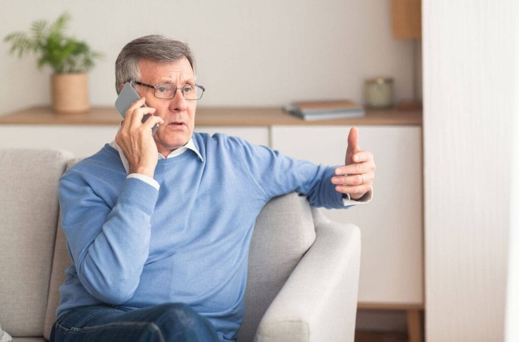 An older adult man sits on a couch talking on a cell phone and looking serious.