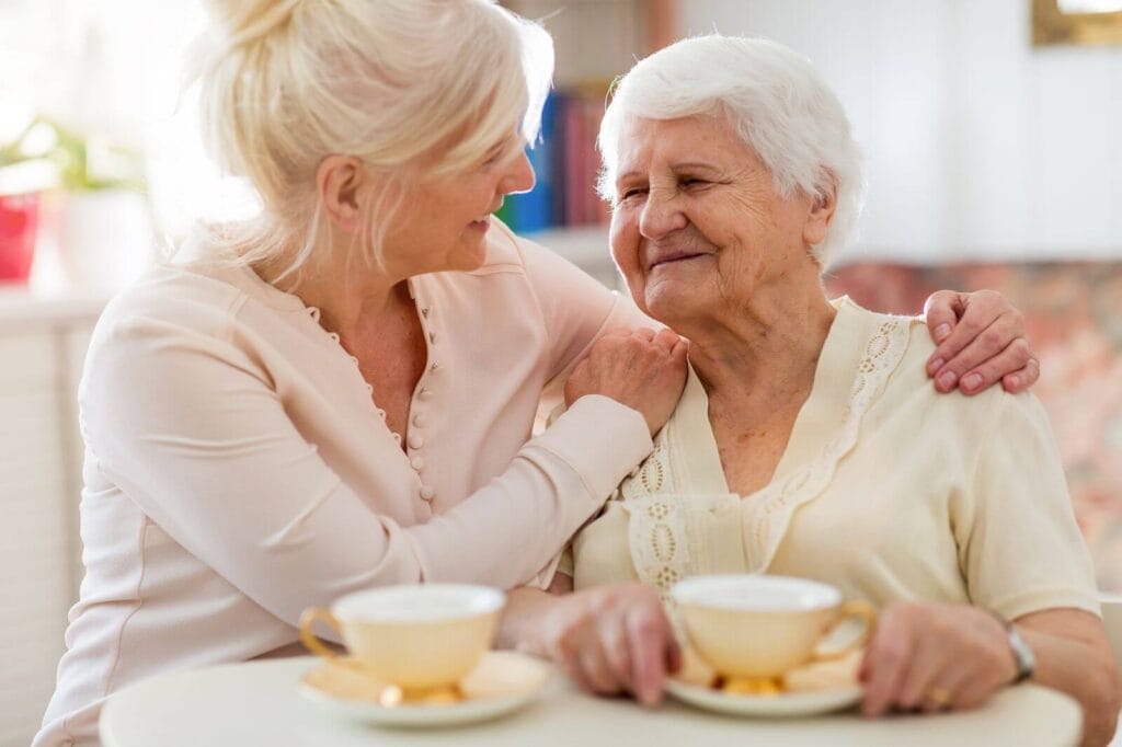An older adult woman and her adult daughter sit at a table drinking tea. The daughter has her arms around her mother's shoulders.