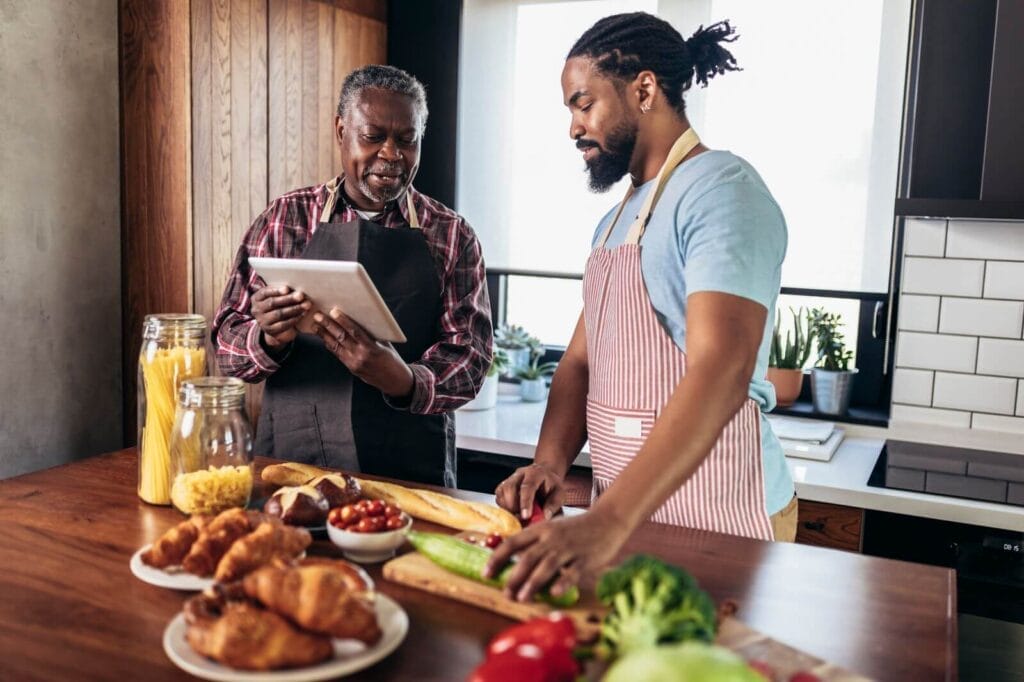 An older adult and his adult son wearing aprons stand at a kitchen island, preparing a meal while consulting a computer tablet.