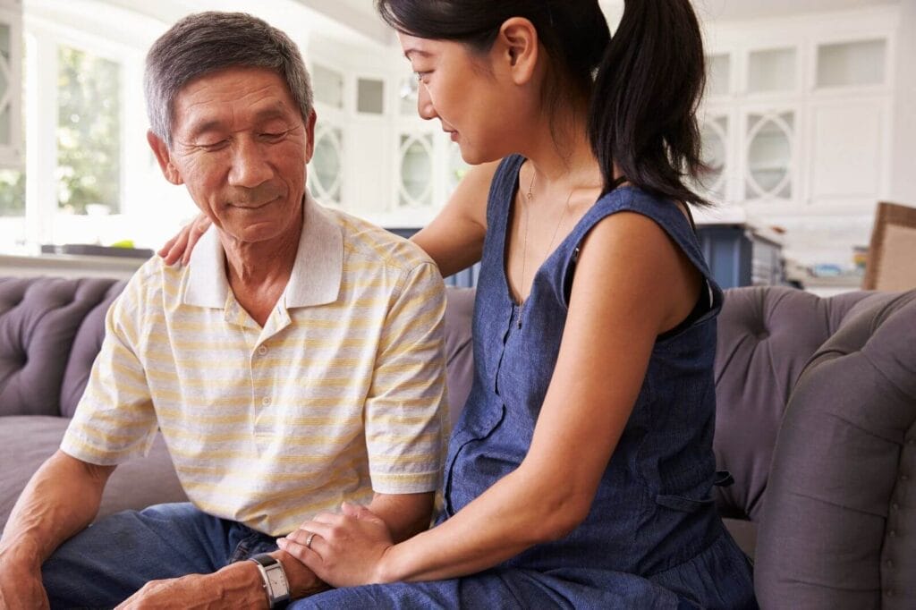 A woman sits on the couch with an older adult man with her arm around his shoulders and her hand on his arm.