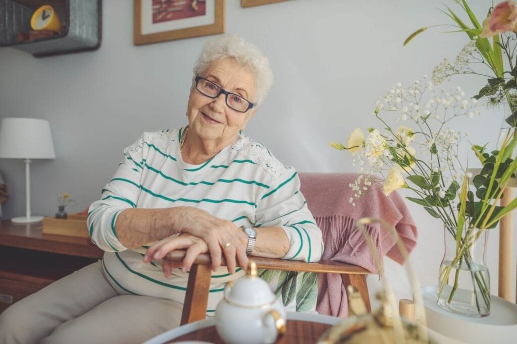 An older adult woman sits in a chair smiling at the camera. There are flowers in a vase next to her and a teapot in front of her. There are pictures hanging on the wall behind her.