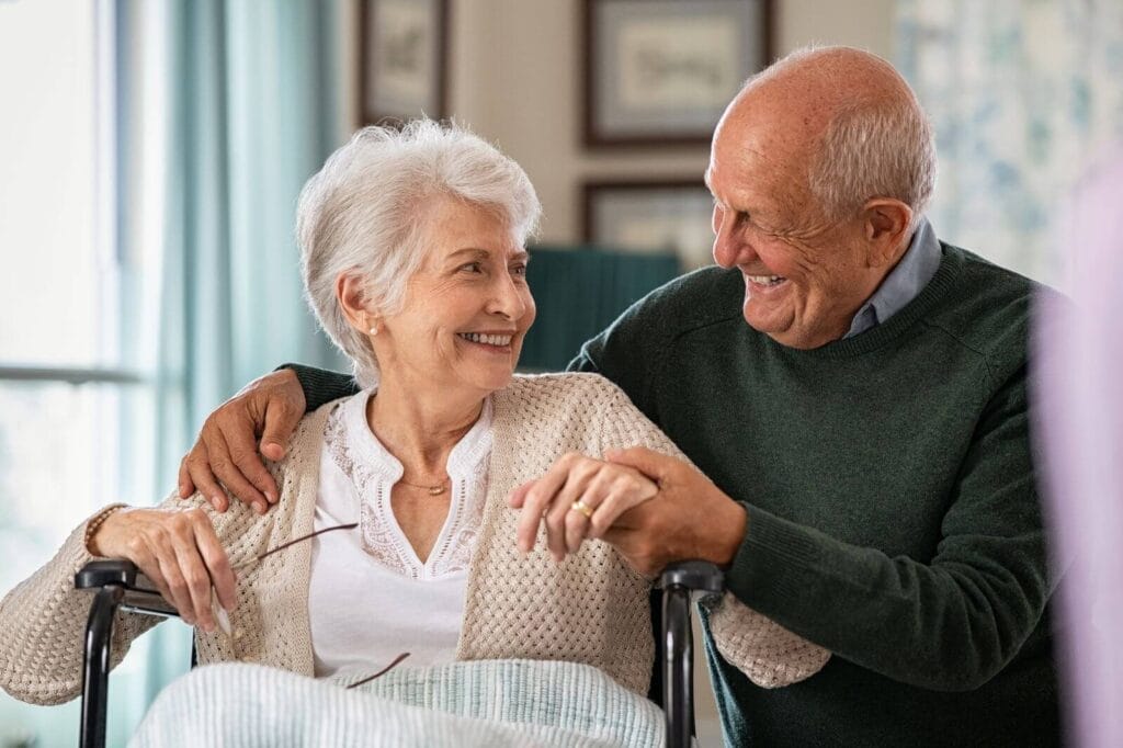 An older adult woman in a wheelchair smiles at her husband, who has his arm around her shoulder and is holding her hand.