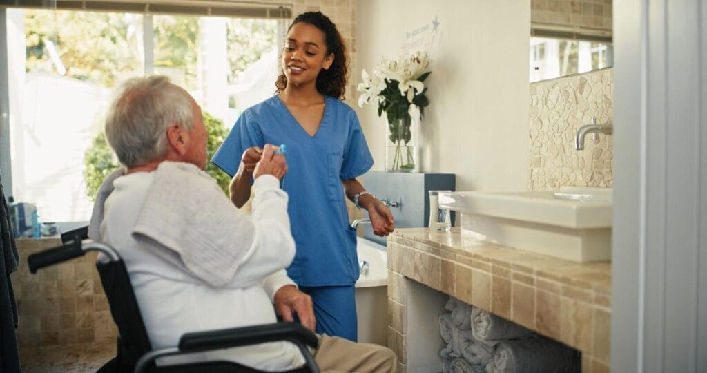 A young woman wearing scrubs hands a toothbrush to an older adult man sitting in a wheelchair.