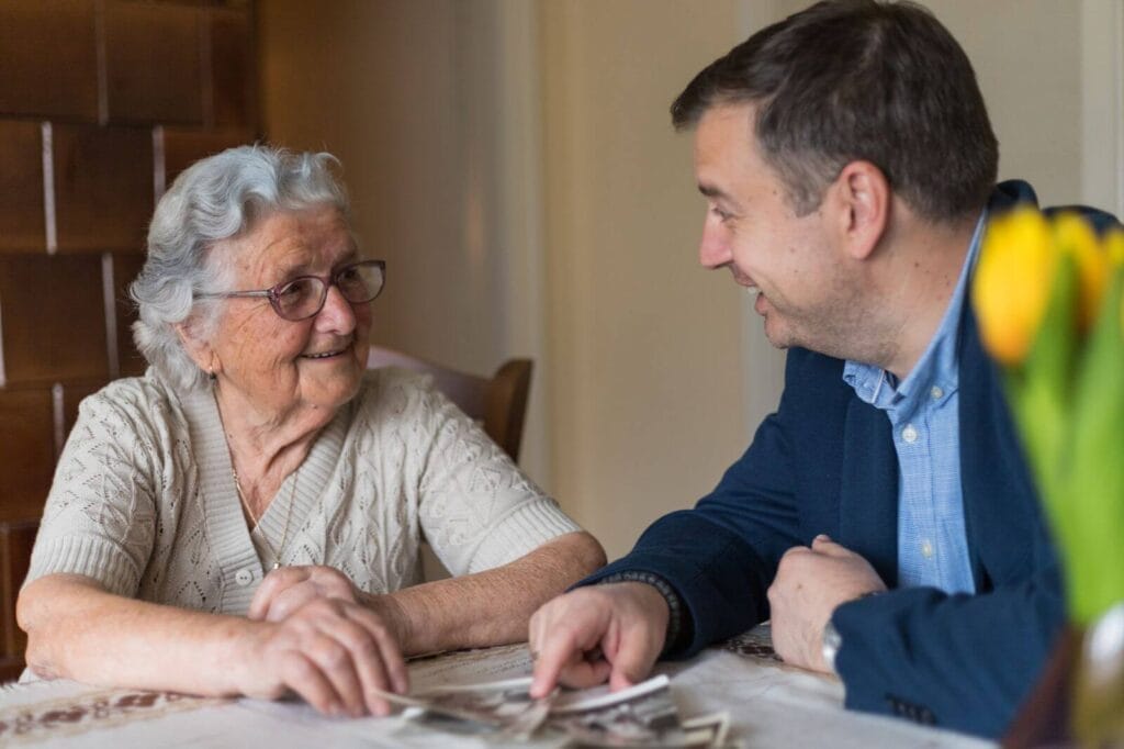 An older adult woman and her son sit at a table looking through old photographs.