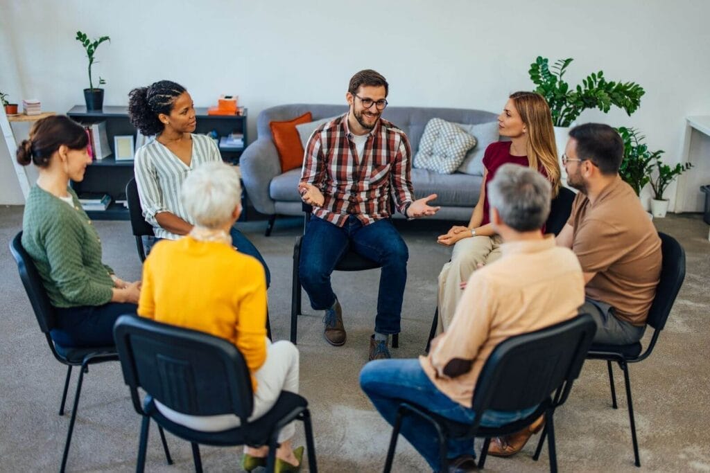 Seven adults of various ages sit in chairs in a circle.