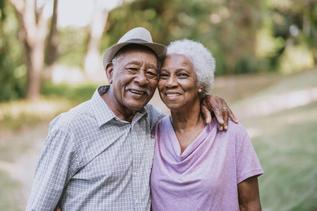 An older adult couple with their arms around each other smile at the camera.