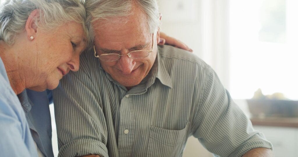 An older adult woman has her arm around the shoulders of an older adult man. Their heads are together.